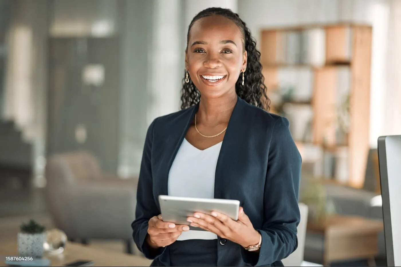 A smiling businesswoman in a blazer, holding a tablet in a bright office.