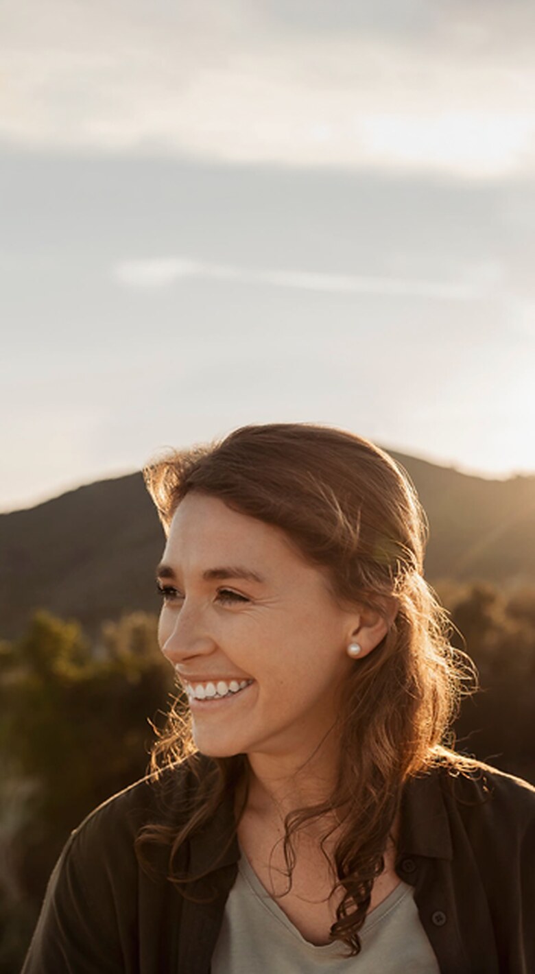 A woman smiling outdoors at golden hour, hills in the background.