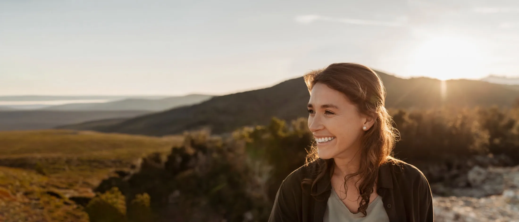 A woman smiling outdoors at golden hour, hills in the background.