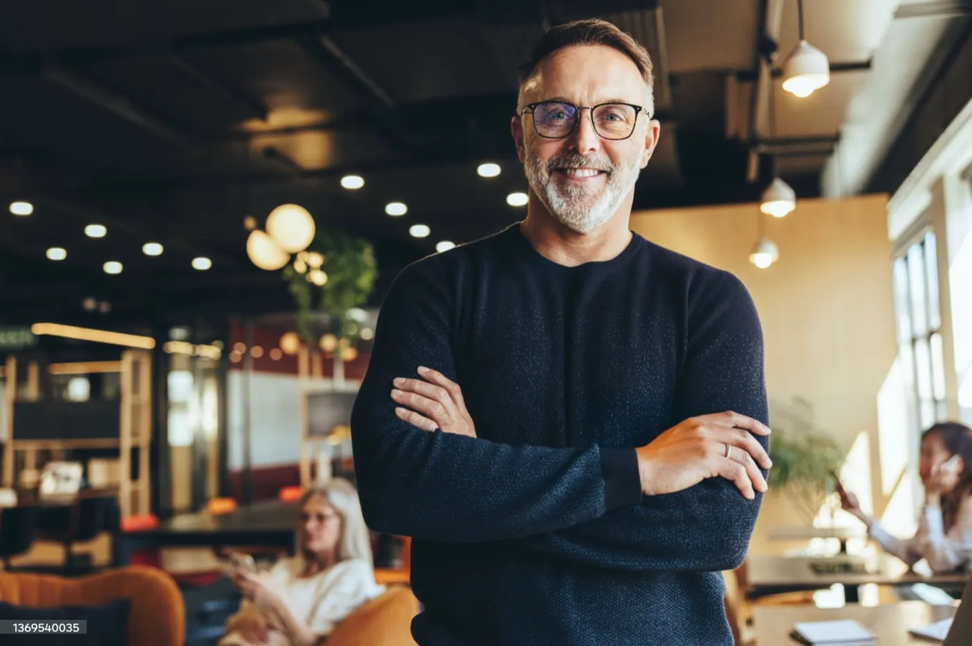 A smiling man with glasses, arms crossed, in a bright indoor space.