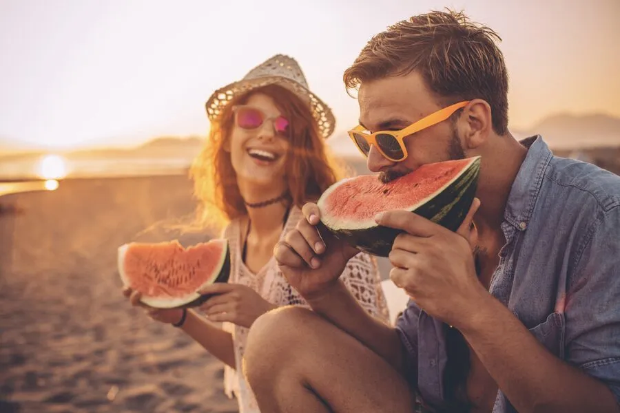 A couple laughing and eating watermelon on the beach.