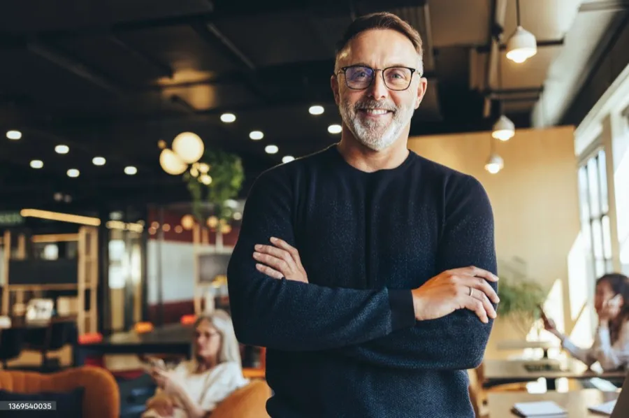 A man with glasses smiling with arms crossed in a cafe.