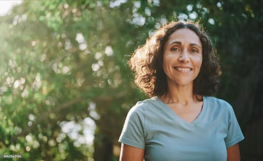 A woman smiling outdoors, dappled light through trees.